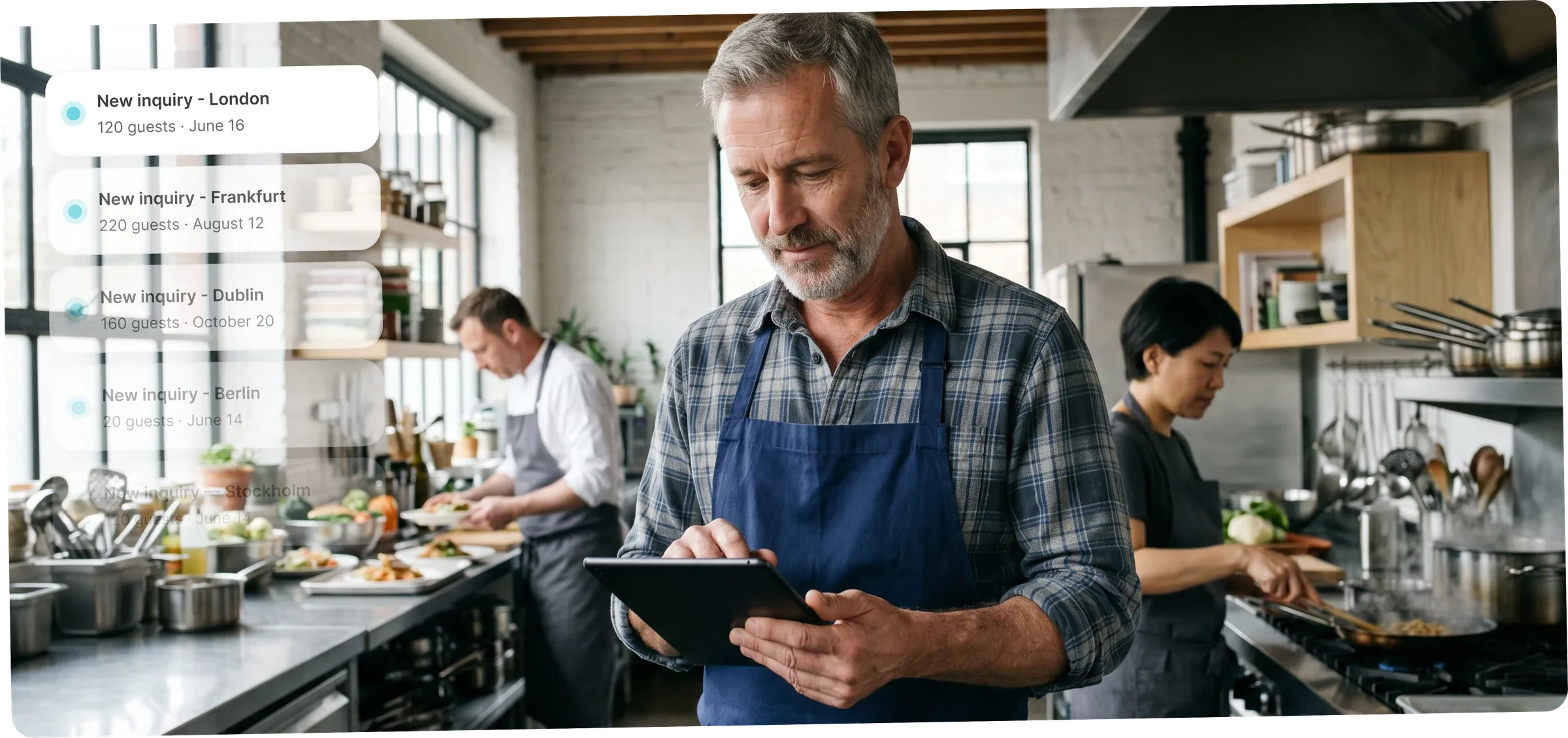 A catering owner with a tablet in a professional kitchen reviews new inquiries in the catermink system; two chefs prepare food in the background